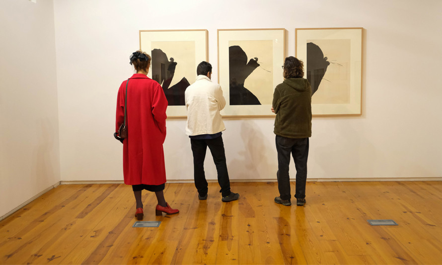 Sala de exposição com três pessoas de costas, olhando para três quadros