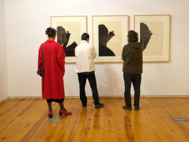 Sala de exposição com três pessoas de costas, olhando para três quadros